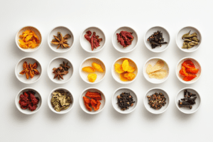 Top-down view of a collection of assorted dried spices and fruits in white bowls, including star anise, dried mango, chili peppers, cloves, and more, arranged neatly on a white background.